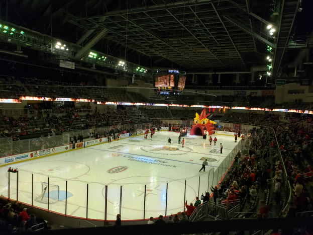 Ein Hockey-Spiel im Columbus Blue Jackets Arena, mit Spielern auf dem Eis, Zuschauern stehend und sitzend, einem umliegenden Zaun, Bannern mit Text, einem Dach mit Metallstangen und Deckenleuchten sowie einem Anzeigebildschirm oben.