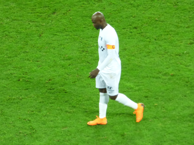 Ein Fussballspieler in weißem T-Shirt, weißen Shorts und orangefarbenen Schuhen überschreitet ein saftig grünes Feld.