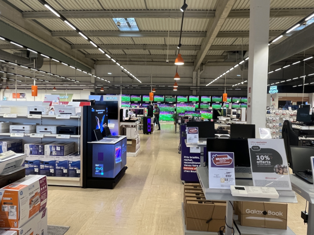 Ein großer Raum in einem Walmart-Laden voller Elektronik, die in Regalen angeordnet ist, einschließlich Computer und Drucker, mit Menschen auf dem Boden, Schildern, Deckenleuchten und Ventilatoren.