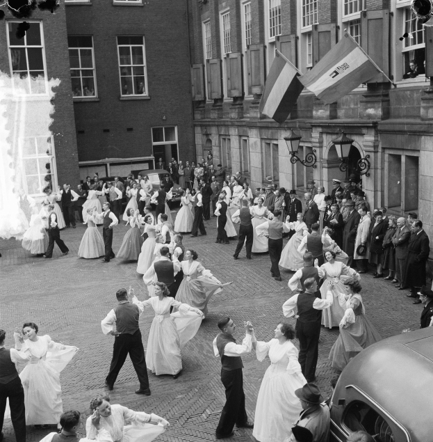 Eine Gruppe von Menschen, die auf der Straße vor einem Gebäude tanzen, mit einem geparkten Auto auf der rechten Seite, Flaggen und Lichter an der Wand des Gebäudes.