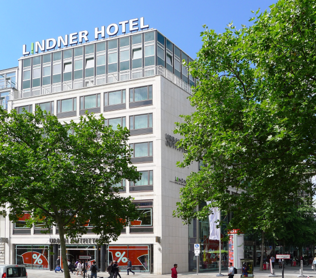 Das Lindner Hotel in Berlin, Deutschland, ein große Gebäude mit Glasfenstern und Text auf der Oberseite, umgeben von Bäumen und Fahrzeugen auf der Straße, mit vielen Menschen, die herumlaufen, einige tragen Taschen, und der Himmel ist im Hintergrund zu sehen.