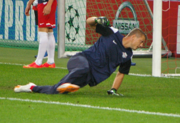 Ein Fußballspieler in Handschuhen taucht auf, um einen Ball vor einem Tor zu fangen, mit zwei Personen, einem Netz und einer Banner mit Text im Hintergrund.