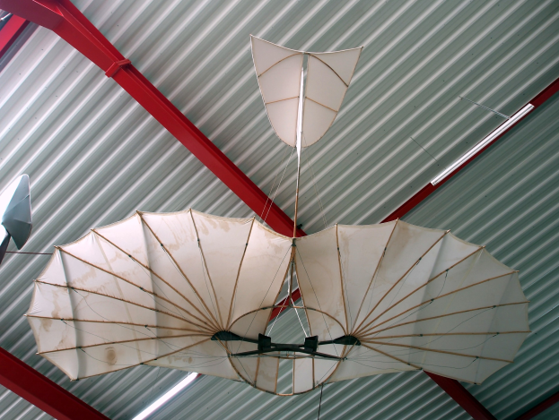 Ein Drachenflieger mit ausgebreiteten Flügeln und rotierenden Propellern hängt von der Metalldecke eines Gebäudes herab bei strahlendem Sonnenschein.