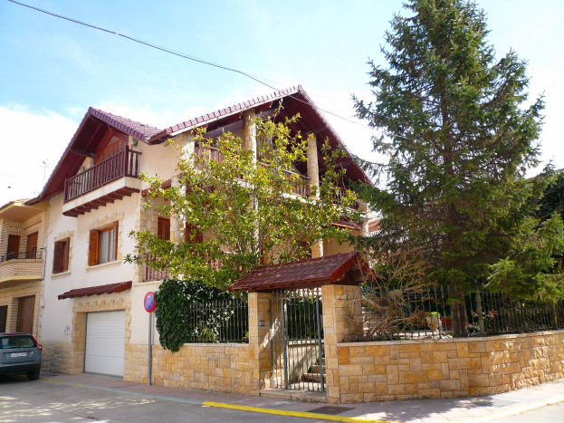 Eine Villa mit einem davor geparkten Auto, mit Fenstern, Geländern, Säulen und einem Dach, umgeben von einem Tor, einem Schild, einem Straßenpfahl, Bäumen und einer Wand, unter einem bewölkten Himmel.