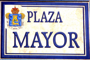 Rechtwinkliges weißes Schild mit schwarzer Schrift "Plaza Mayor" in fetter Schrift, blauer Rahmen und gekröntem Wappen-Logo an einer Gebäudefassade.