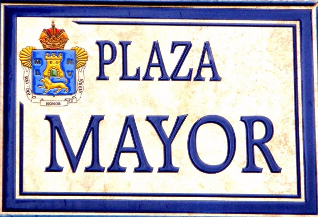 Rechtwinkliges weißes Schild mit schwarzer Schrift "Plaza Mayor" in fetter Schrift, blauer Rahmen und gekröntem Wappen-Logo an einer Gebäudefassade.