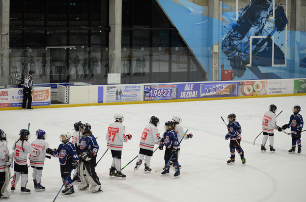 Junge Menschen in Helmen und Hockey-Ausrüstung spielen Eis-hockey auf einer Indoor-Eisbahn mit Bannern, Säulen und Glaswänden im Hintergrund.