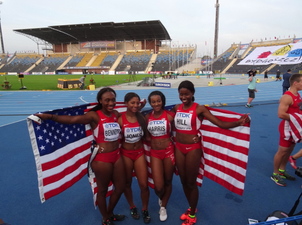 Vier Frauen in Sportkleidung stehen zusammen auf einer Laufbahn, lächeln und halten amerikanische Flaggen, mit einem Stadion und Himmel im Hintergrund.