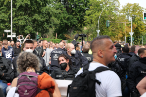 Eine große Gruppe von Menschen, einige mit Masken und Rucksäcken oder Kameras, steht vor einer Menge von Polizeibeamten, mit Laternenmasten, Ampeln, Verkehrsschildern, Bäumen und einem klaren blauen Himmel im Hintergrund.