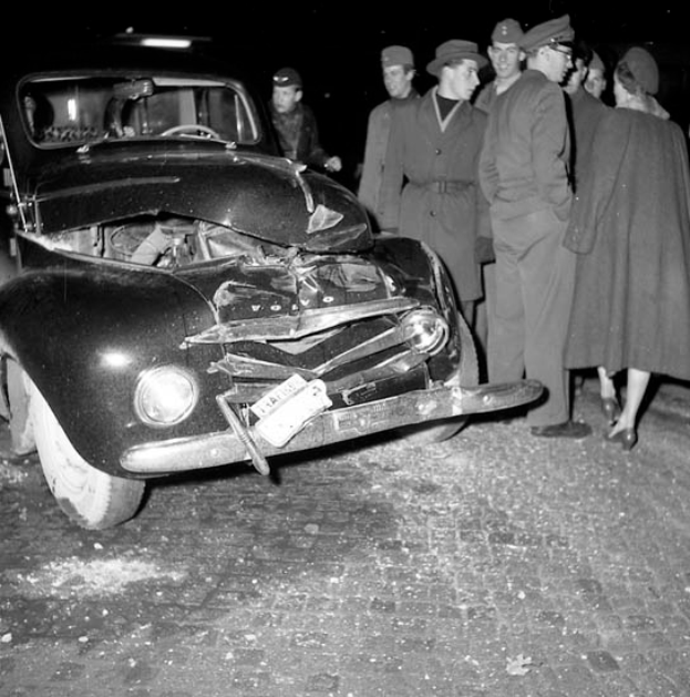 Ein Schwarz-Weiß-Bild eines beschädigten Autos am Straßenrand, mit einer Gruppe von Menschen drumherum, die das Auto besorgt betrachten.