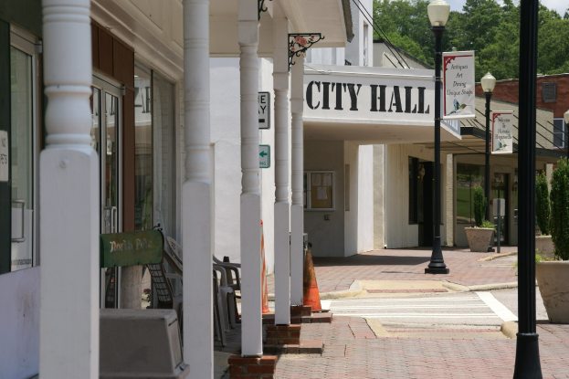 Ein Rathausturm mit Gebäuden, Straßenmasten, Straßenlaternen, Namensschildern, Kraftfahrzeugen auf der Straße, Absperrgittern, Stühlen, Topfpflanzen, Bäumen und einem bewölkten Himmel im Hintergrund.