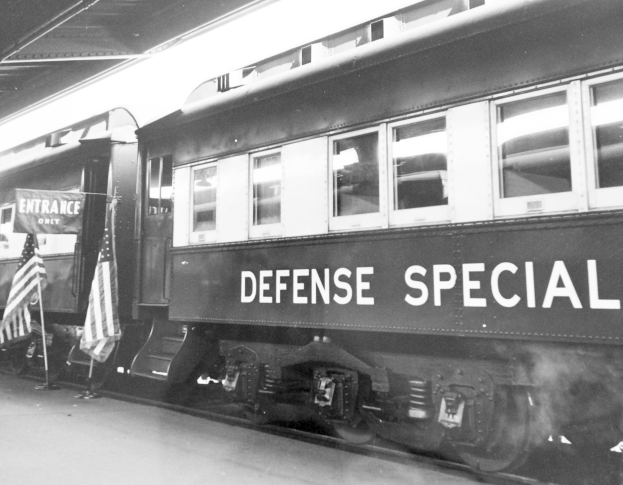 Schwarz-weiß-Foto eines Zuges mit der Aufschrift "Defense Special" auf den Gleisen mit zwei Fahnen links und einer Decke darüber.