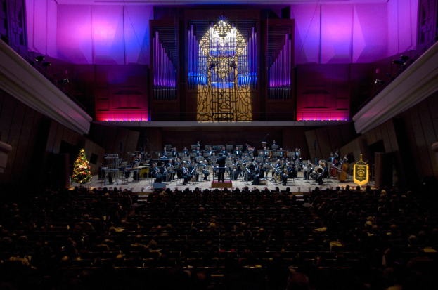 Ein großes Auditorium voller Menschen bei einem Konzert, Musiker auf der Bühne, ein Weihnachtsbaum im Hintergrund und Wände mit Lichtern und anderen Gegenständen geschmückt.