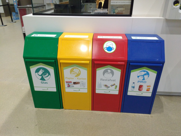 Vier bunte Mülltonnen in einer Reihe auf dem Boden vor einer Theke mit einem Monitor und anderen Gegenständen, mit einer teilweise sichtbaren Person auf der linken Seite und an der Wand befestigten Gegenständen auf der rechten Seite.