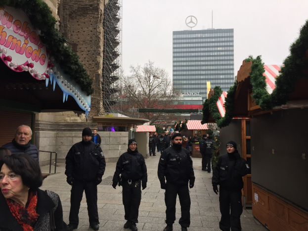Eine Gruppe von Polizisten in schwarzen Uniformen und Mützen steht vor einem geschmütckten Weihnachtsmarkt in Berlin, mit festlichen Dekoelementen, Gebäuden, Bäumen und einem klaren blauen Himmel im Hintergrund; ein Schild mit Text ist links sichtbar.