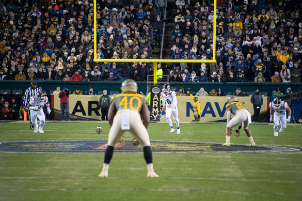 Ein Fussballspiel wird in einem Stadion mit Spielern in Sportkleidung und Helmen im Vordergrund und einer Zuschauermenge im Hintergrund sowie sichtbaren Pfählen und Tafeln mit Text gespielt.