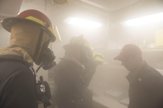 Feuerwehrleute in einem raucherfüllten Raum während einer Übung, einer hält eine Kamera, mit Hintergrundbeleuchtung und -ausrüstung