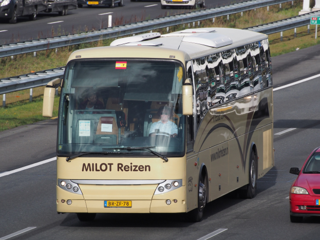 Ein Minibus mit sichtbaren Insassen auf einer Autobahn neben einem roten Auto fahrend, mit Geländern und Gras auf beiden Seiten der Straße und Texttafeln auf Säulen im Hintergrund.