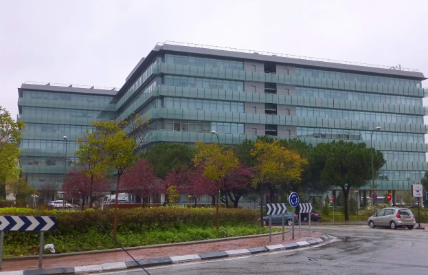 Großes modernes Gebäude mit zahlreichen Fenstern, identifiziert als Europäisches Zentralbank-Hauptquartier, umgeben von Grünflächen, Straßeninfrastruktur und Fahrzeugen unter einem bewölkten Himmel.
