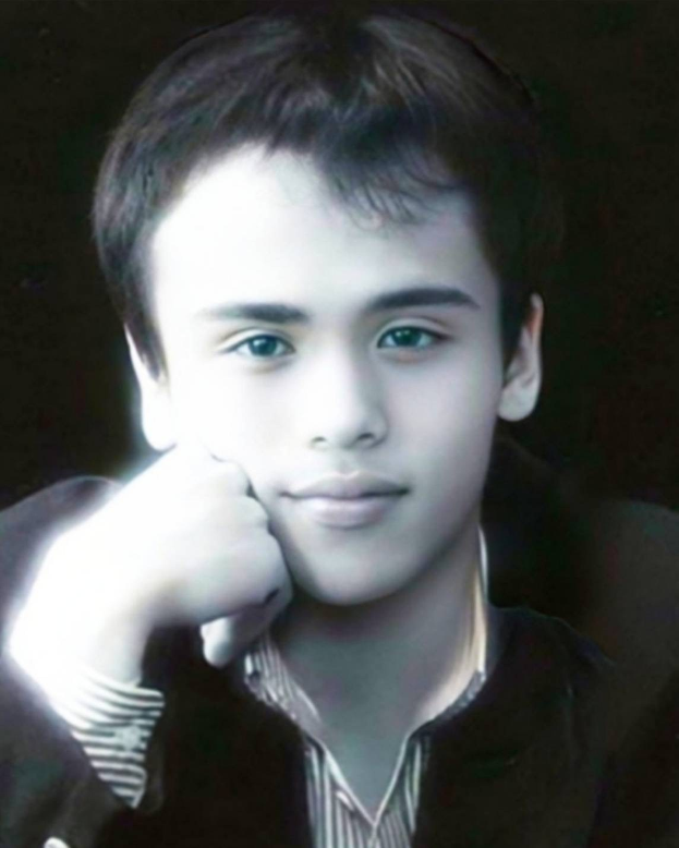 Ein junger Mann in einer schwarzen Jacke posiert selbstbewusst vor einem dunklen Hintergrund mit einem entschlossenen Gesichtsausdruck und leuchtenden Augen.