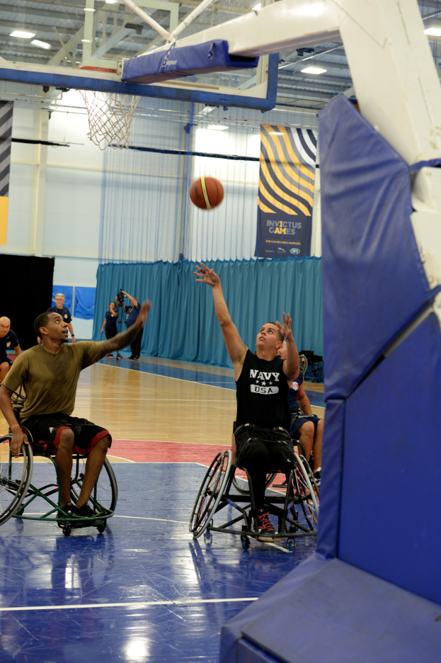 Eine Gruppe von Menschen in Rollstühlen spielt Basketball in einer Turnhalle mit einem Basketballkorb, Bannern mit Text und Deckenleuchten.