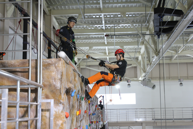 Zwei Personen in schwarzen und orangen Outfits und Helmen klettern an einer Felswand in einer Halle, gesichert durch Seile, mit einem Metallgeländer links und einer weißen Wand mit Leuchten im Hintergrund.