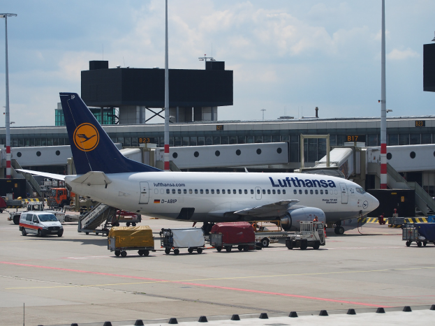 Lufthansa Airbus A320-200 am Frankfurt Airport mit Bodenfahrzeugen und Wagen vorne, einem Gebäude, Laternenmästen und einem klaren blauen Himmel im Hintergrund.