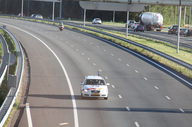 Ein Auto fährt auf einer Autobahn mit einem stationären Polizeiwagen am Straßenrand, andere Fahrzeuge und Bäume, Geländer, Pfosten und Bretter im Hintergrund.