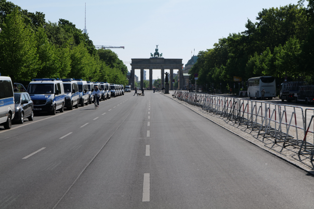 Eine Reihe von Polizeifahrzeugen, die auf einer Straße vor dem Brandenburger Tor in Berlin, Deutschland, geparkt sind, mit Menschen auf Fahrrädern und Stehenden, Barrieren, Bäumen und einem Bogen mit Statuen im Hintergrund.