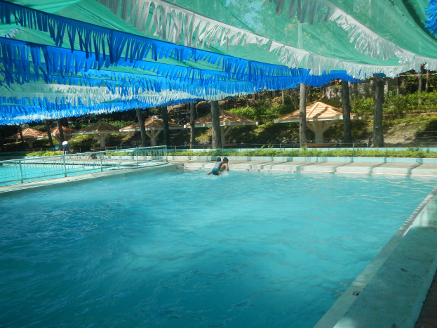 Ein großes Schwimmbad mit schwimmenden Menschen, umgeben von einem Zaun, Pflanzen, Bäumen, Hütten und von der Decke hängenden Dekorationen.