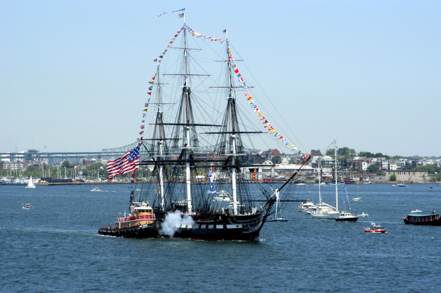 Ein Segelschiff mit Flaggen und Masten segelt nahe an einer Stadt vorbei, umgeben von anderen Booten, mit Gebäuden, Bäumen und einem klaren blauen Himmel im Hintergrund.