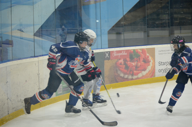 Gruppe junger Menschen, die Eis-Hockey auf einer Indoor-Eisfläche spielen, mit Helmen, Sportkleidung und Hockey-Schlägern, mit einem Plakat im Hintergrund auf einer Glaswand.