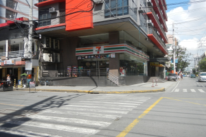 Eine belebte Stadtstraße mit Fahrzeugen, Fußgängern, hohen Gebäuden, einem 7-Eleven-Laden, Strommasten, Bäumen und einem bewölkten Himmel.