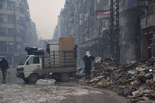 Ein Lastwagen fährt auf einer Straße neben einem Schutthaufen, mit Menschen, die auf der Straße gehen, einer hält einen Schirm, und Gebäuden, Pfählen und Brettern mit Text im Hintergrund unter einem klaren Himmel.