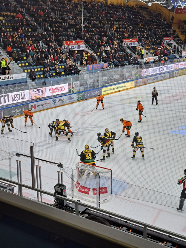 Eine Gruppe von Menschen, die Hockey auf einem Eisplatz in einer Arena spielen, mit einem Netz, einer Umzäunung, sitzenden Zuschauern, Bannern und Scheinwerfern.