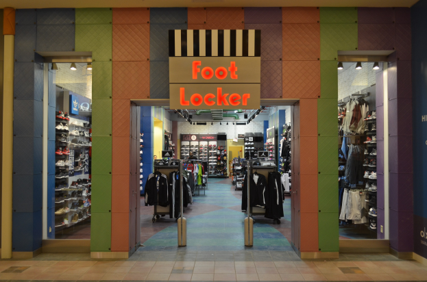 Innenansicht eines Foot Locker Ladens mit Kleidung an Bügeln und Regalen sowie Deckenbeleuchtung und sichtbarem Boden.