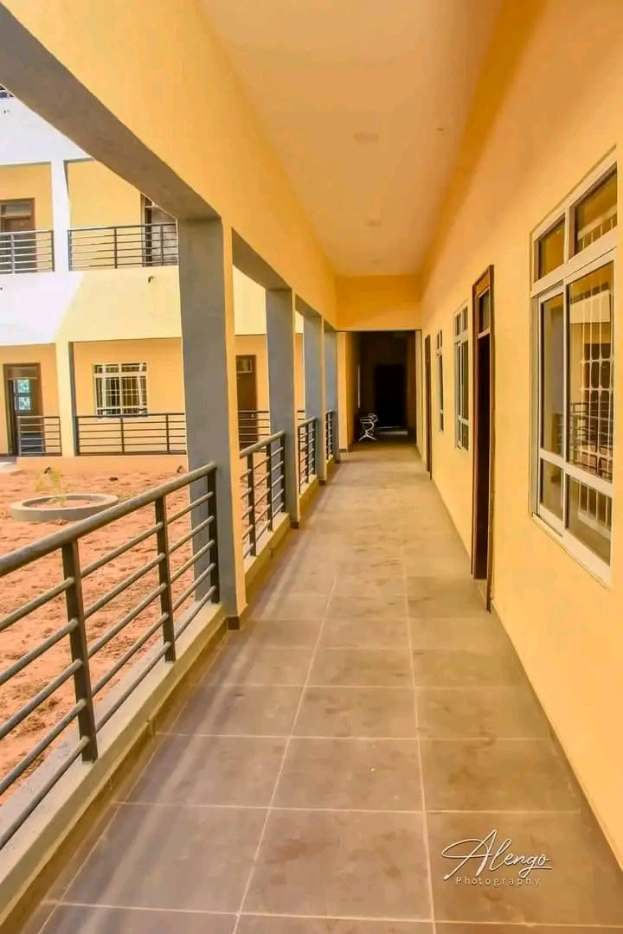 Ein Gebäude mit Fenstern, Türen, Säulen, Geländern und einem langen Flur mit einem gefliesten Boden und einem Balkon, mit anderen Gebäuden und einem klaren blauen Himmel im Hintergrund, und einem Text am unteren Rand, der "3-Zimmer-Wohnung zur Miete in Ntinda, Kampala" liest.