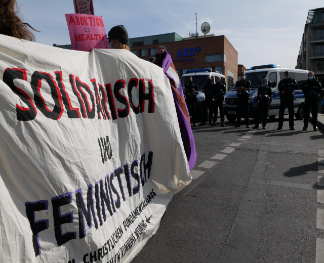 Gruppe von Menschen, die eine "Solidarität und Feminismus"-Fahne schwingend durch die Straße marschieren, mit parkenden Fahrzeugen, Gebäuden, einer Schüsselantenne und einem klaren blauen Himmel im Hintergrund.
