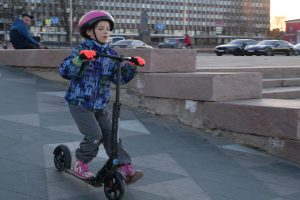 Ein junger Junge fährt mit einem Helm und Handschuhen auf einem Gehweg mit einem E-Scooter, umgeben von verschiedenen städtischen Elementen und einem klaren blauen Himmel.