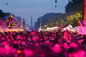 Eine große Menschenmenge auf einem nächtlichen Musikfestival mit farbigen Lichtern, Ballons, Zelten, Bannern, Fahnen, Bäumen, einem Turm und einem sternenklaren Himmel im Hintergrund.