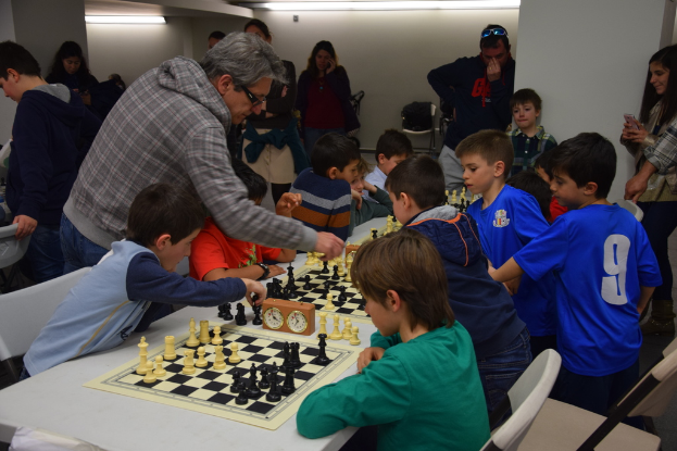 Kinder spielen Schach bei einem Wettbewerb, während Erwachsene aus dem Hintergrund und den Seiten zuschauen.