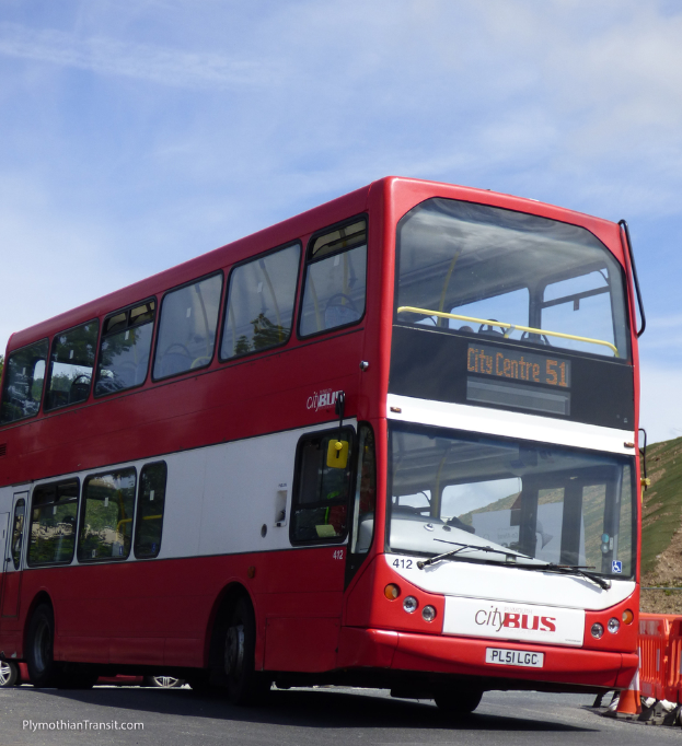 Ein roter Doppeldeckerbus mit der Aufschrift "Stadtbus" fährt auf einer Straße mit einem Verkehrskegel im Vordergrund und einem Hügel im Hintergrund.