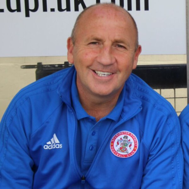 Zwei Männer in einem Raum, einer in einem blauen Jackett, der neben einem anderen Mann lächelt, mit einer Tafel an der Wand dahinter.