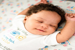 Ein Baby, das auf einer Wickelauflage schläft und ein weißes T-Shirt trägt, auf dem 'Sleep Safe for Kids' steht, mit einem unscharfen Hintergrund.