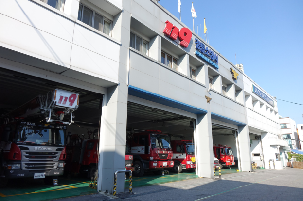 Feuerwache mit mehreren Feuerwehrautos vor dem Gebäude geparkt, mit Fenstern, Säulen, Namensschildern und Fahnenmasten im Vordergrund, vor anderen Gebäuden unter einem klaren blauen Himmel.