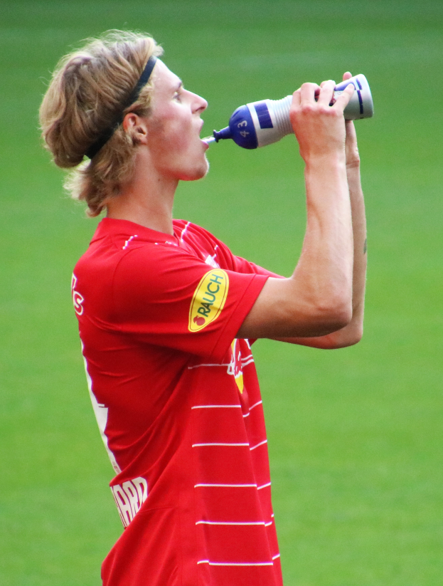 Ein Mann in rotem T-Shirt trinkt aus einer Flasche, während er auf einem Fussballfeld steht.