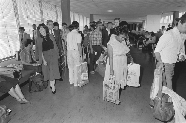 Schwarzes und weißes Bild von Reisenden in einem Flughafen, einige gehen mit Gepäck, andere sitzen auf Bänken oder stehen, mit einem Mann und einer Frau in der Mitte, die Taschen halten.