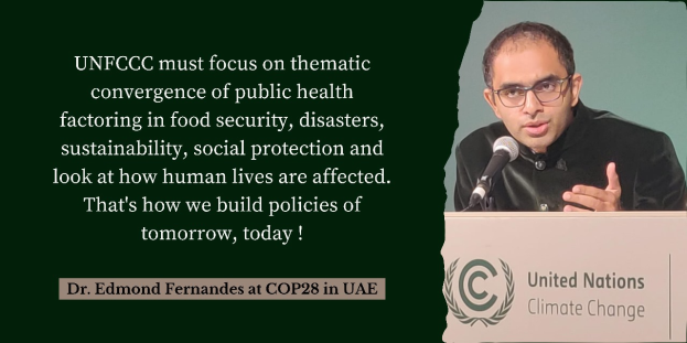 Dr. Edmund Fernandes, der eine schwarze Robe und eine Brille trägt, spricht in ein Mikrofon auf der COP28 in den VAE, vor einem Pult mit einem Logo und Text, während der Text auf der linken Seite "UNFCC muss sich auf die thematische Zusammenführung von Gesundheit, Lebensmittelsicherheit, Katastrophen, Nachhaltigkeit, sozialem Schutz konzentrieren und darauf achten, wie das Leben der Menschen beeinflusst wird."