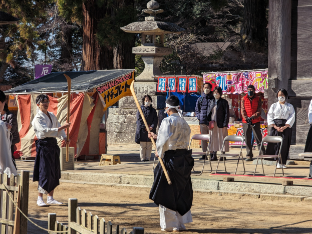 Eine Gruppe von Menschen in traditioneller Kleidung steht äußerlich in Kyoto, einige tragen Masken und halten hölzerne Stäbe, mit Stühlen, Bannern und einem Zelt im Hintergrund unter einem klaren blauen Himmel.