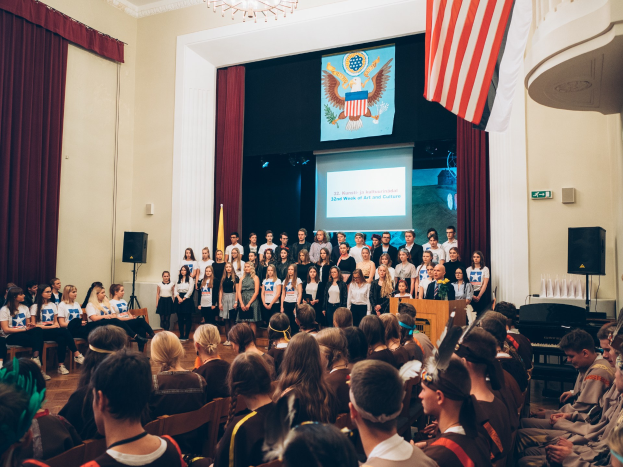 Eine Gruppe von Menschen steht an einem Podium mit einem Mikrofon, flankiert von Flaggen und Lautsprechern, die eine sitzende Menge während eines kulturellen Ereignisses ansprechen, mit einem Bildschirm, Vorhängen und einem Kronleuchter im Hintergrund.